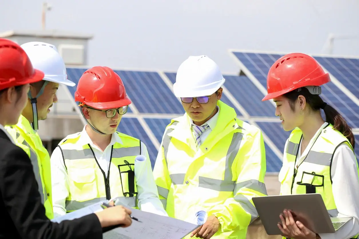 Three men in hard hats and safety vests are standing near a solar panel.